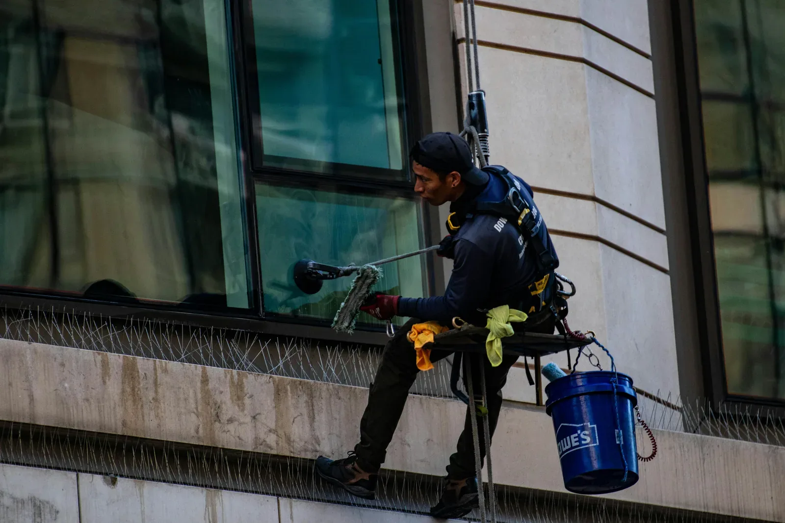 Glasreiniger im Sicherheitsgeschirr putzt mit Wischer und Eimer eine Fensterfront an einer Gebäudefassade